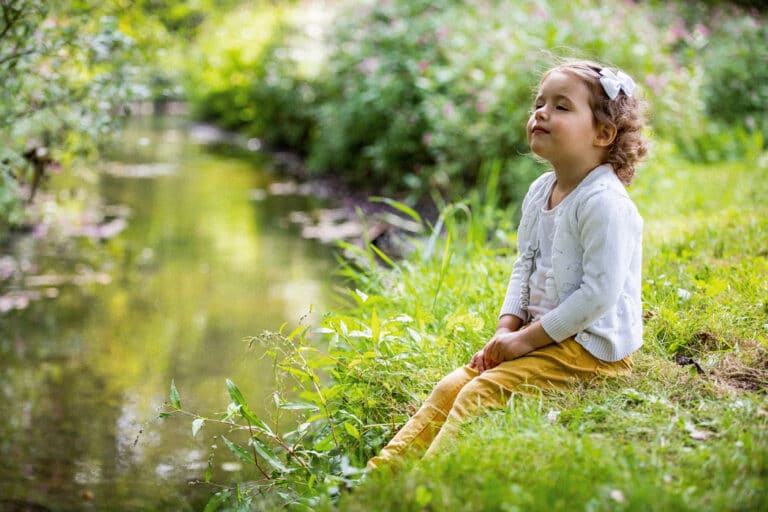 Ätherische Öle für Kinder: Kind sitzt in einer Wiese vor einem Fluss und riecht die Natur