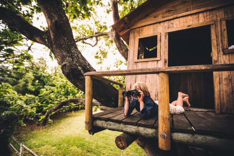 Baumhaus: Kind mit Fernglas liegt auf dem Bauch im Baumhaus